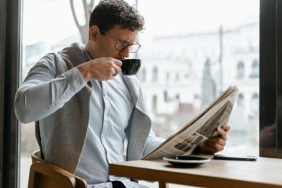 Man drinking coffee while reading a newspaper at a café window seat, reflecting on real estate market news